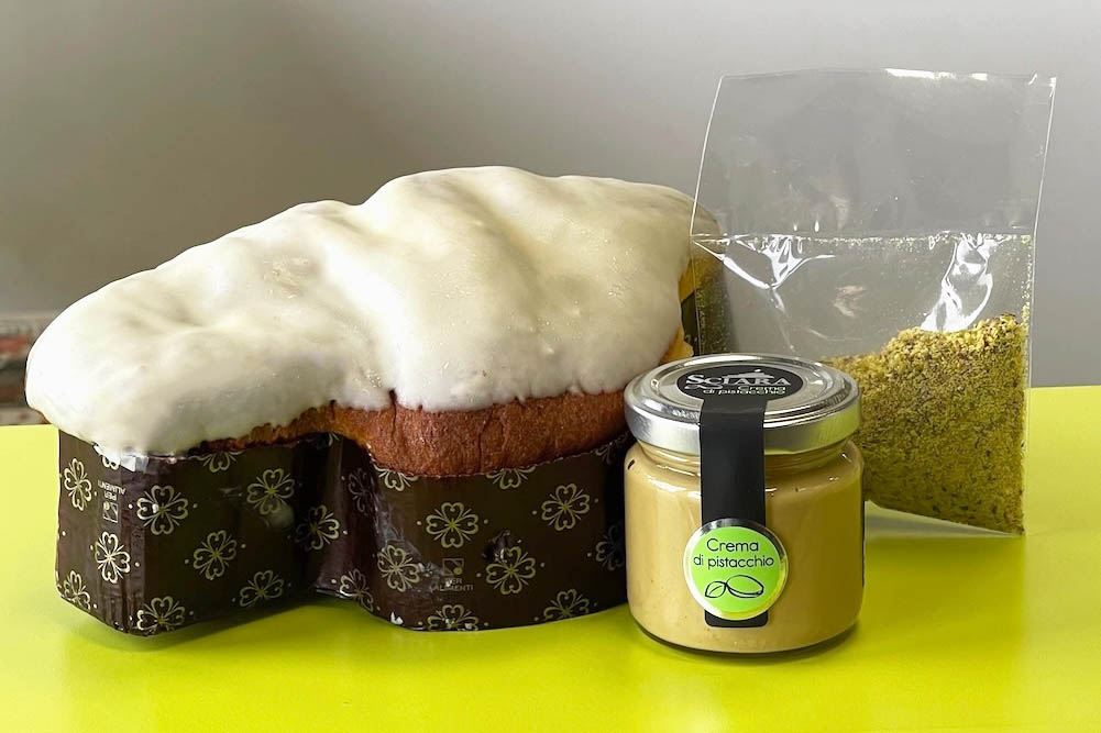Glutenfreie Pistazien-Colomba von Sciara mit Pistaziengranulat oben drauf und einem Glas Sahne an der Seite.